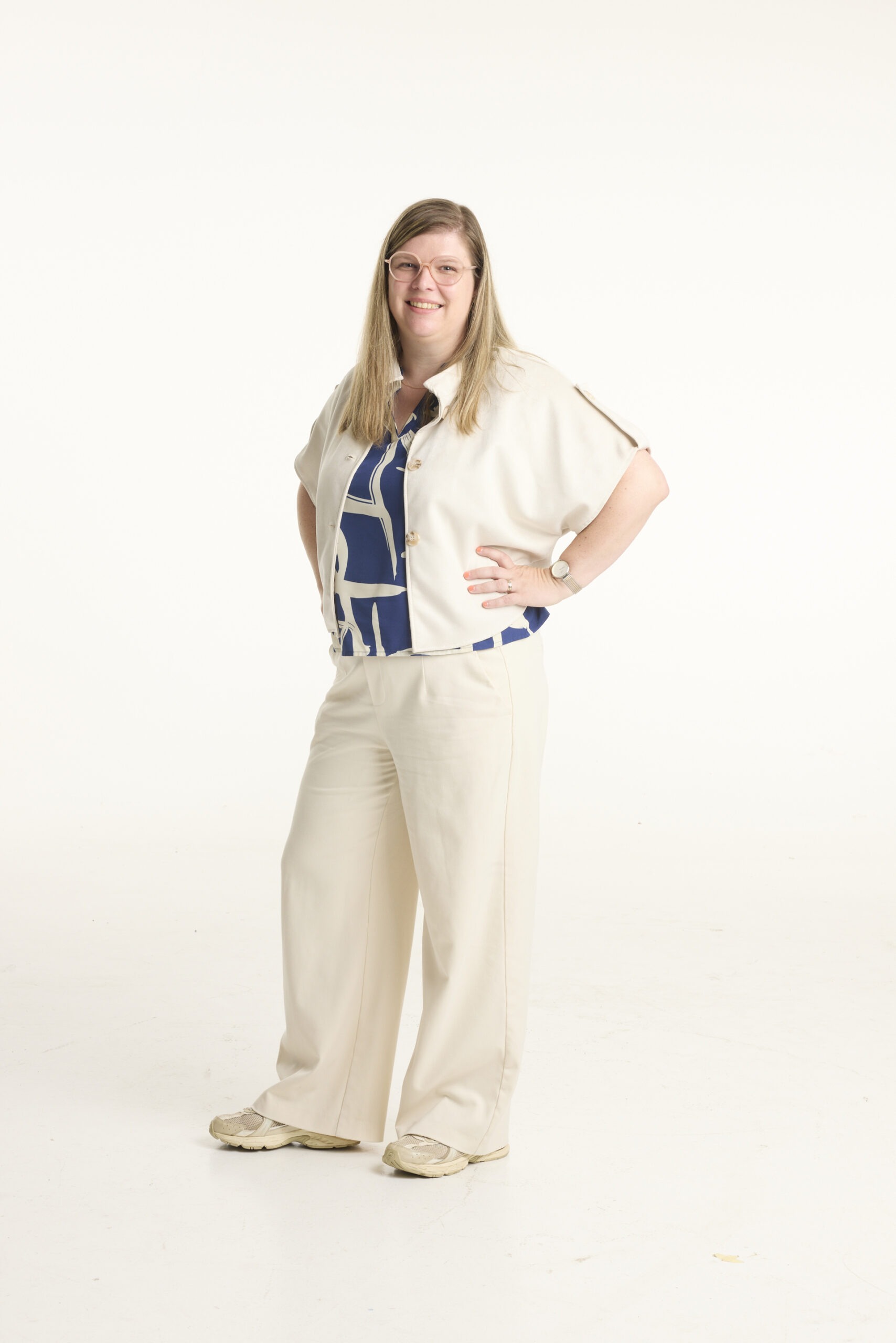 Woman standing in beige outfit with hands on hips, smiling at camera, wearing glasses and a blue patterned blouse against a light studio backdrop.