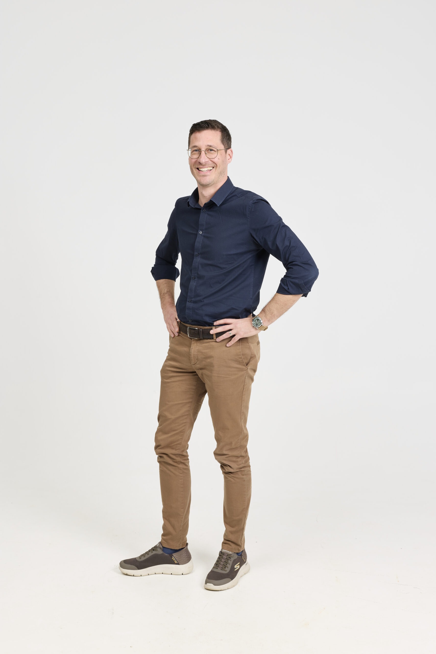 Smiling man in a navy shirt and tan pants stands with hands on hips against a light background, wearing glasses and sneakers.