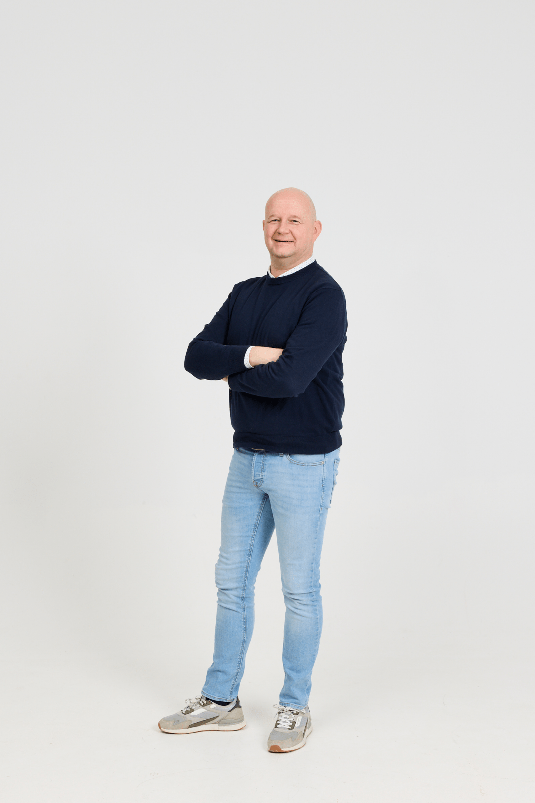 Man standing with arms crossed, smiling at the camera against a plain white studio background, wearing a navy sweater and light jeans.